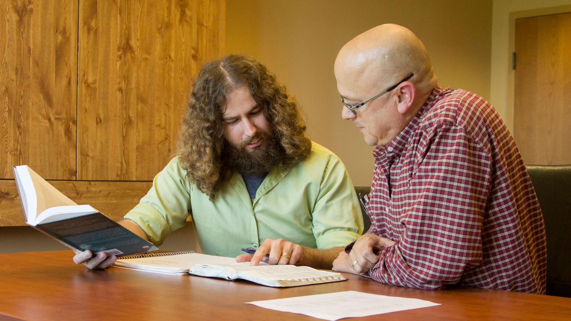 professor and student sit at table looking at Bible and discussing it with each other