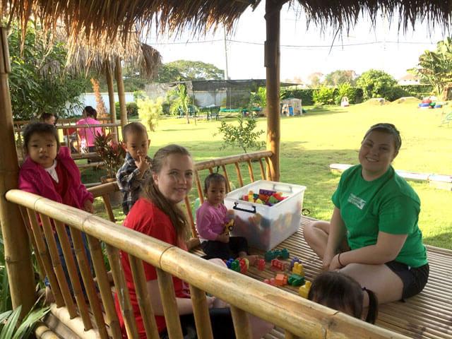 two female 学生 smile while playing with young children in Thailand